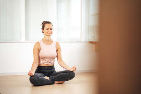 Full length of Caucasian young woman practicing yoga lotus position in apartmentの写真素材
