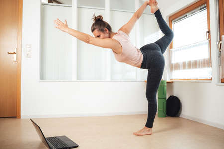 Fit young woman standing on one leg practicing yoga while watching laptop at homeの写真素材