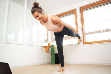 Full length of young sporty woman watching laptop while balancing on one leg at homeの写真素材