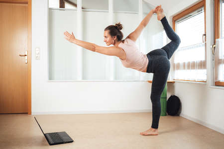 Full length of sporty young woman standing on one leg practicing yoga with laptop at homeの写真素材