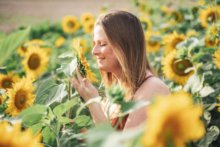 Smiling Caucasian woman with blond hair smelling sunflower in farm on sunny dayの写真素材