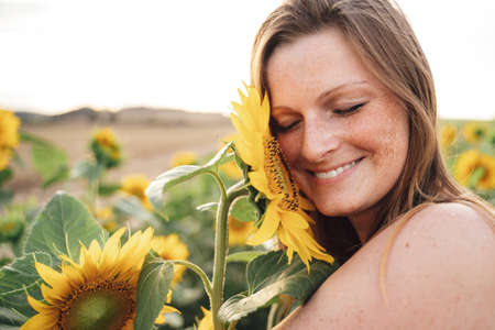 Close up of smiling young woman with eyes closed touching sunflower on face at fieldの写真素材