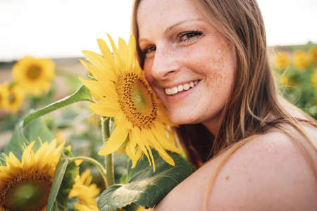 Close up portrait of smiling young woman with fresh yellow sunflower at farmの写真素材