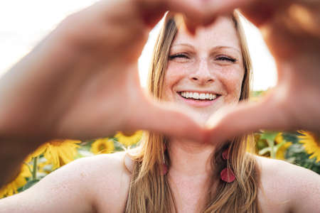 Close up portrait of smiling young woman making heart shape with her hands at sunflower fieldの写真素材