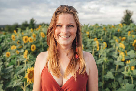 Portrait of smiling young woman with long blond hair with sunflower plants in background in sunny dayの写真素材