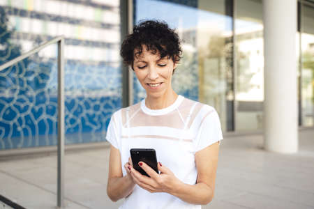 Mature businesswoman with short black hair using mobile phone outside office building in cityの写真素材