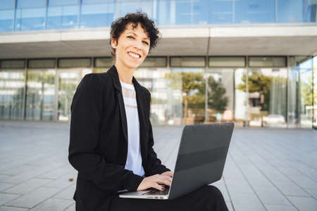 Portrait of smiling mature businesswoman in suit using laptop while sitting outside officeの写真素材