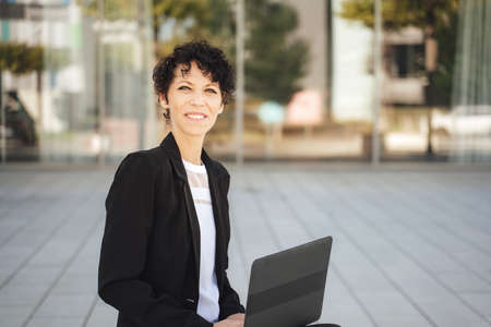 Portrait of smiling Caucasian businesswoman in suit sitting with laptop outside officeの写真素材