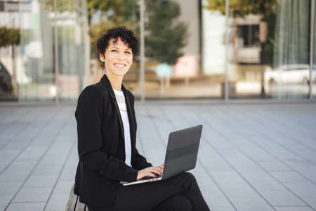 Portrait of smiling mature businesswoman working on laptop outside near officeの写真素材