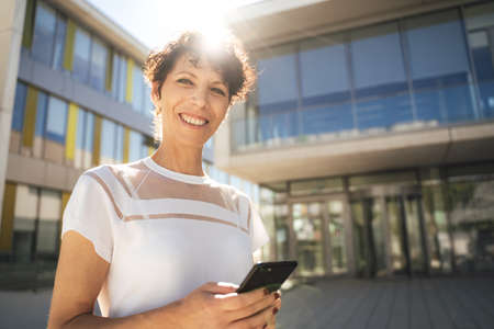 Portrait of smiling mature female freelancer holding mobile phone in city on sunny dayの写真素材