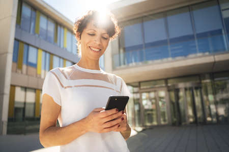 Smiling mature woman using mobile phone outside building on sunny day in cityの写真素材