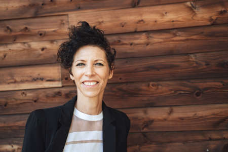 Close up portrait of smiling mature businesswoman in suit standing against wooden wallの写真素材