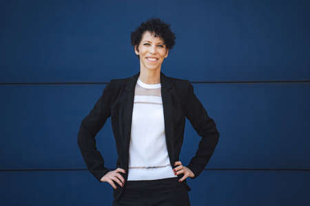Portrait of smiling mature businesswoman standing with hands on hip against blue wallの写真素材