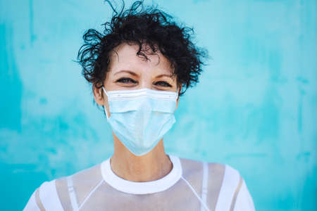 Close up portrait of mature woman with curly short hair wearing safety mask against blue wallの写真素材