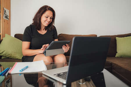 Smiling mid adult female freelancer using digital tablet while sitting on sofa at homeの写真素材