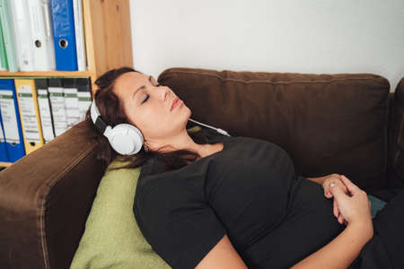Beautiful Caucasian woman lying on sofa while listening music through headphones at homeの写真素材