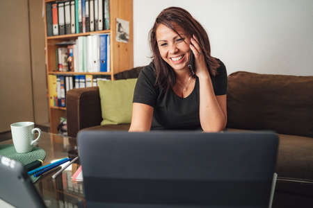 Smiling businesswoman talking on smartphone while using laptop in living room at homeの写真素材