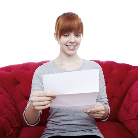 young beautiful red haired girl on red sofa reading a letter in front of white backgroundの写真素材
