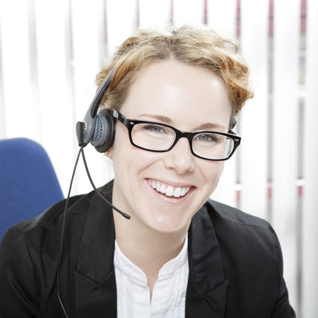 Happy enthusiastic young business woman with a beautiful friendly smile wearing a headset and glasses sitting in an office looking at the cameraの写真素材