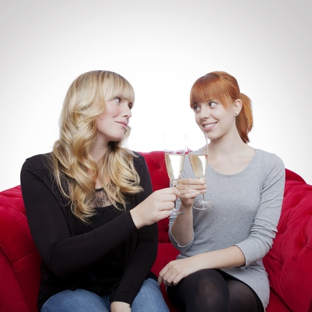 young beautiful blond and red haired girls with champagne on red sofa in front of grey backgroundの写真素材