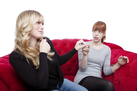 young beautiful blond and red haired girls get cigarette away on red sofa in front of white backgroundの写真素材