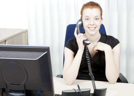 Smiling happy business woman, personal assistant or secretary sitting behind her desk with a telephone handset in her handの写真素材