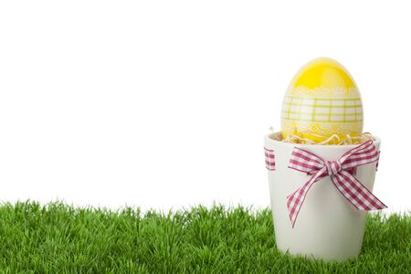Pretty Easter gift with a decorative yellow Easter Egg inside a flower pot tied with a rustic red and white checked ribbon and bow standing on green grass against a white background with copyspaceの写真素材
