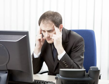 Worried businessman with a headache sitting at his desk rubbing his temples with his fingertips and staring at his desktop computer monitor in despairの写真素材
