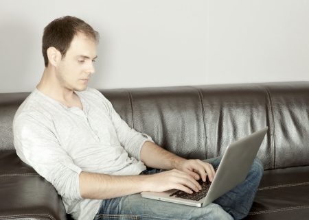 Attractive middle-aged man relaxing on a large black leather sofa at home typing on his laptop as he conducts his business or surfs the internet entering dataの写真素材
