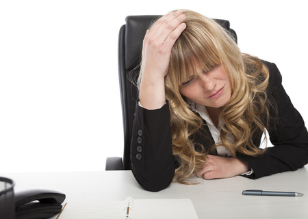 Worried businesswoman with long blond hair sitting at her desk with her hand on her head staring at the desktop as she sits thinkingの写真素材