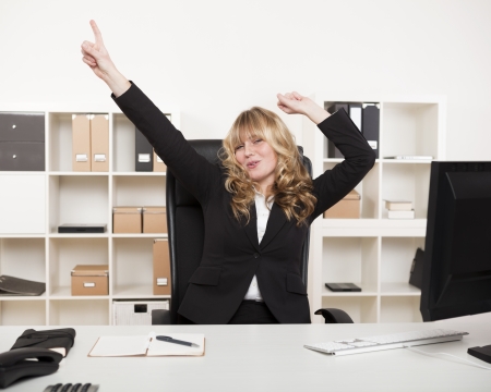 Jubilant businesswoman celebrating a success pointing and punching the air with her fist as she sits at ger desk in the officeの写真素材
