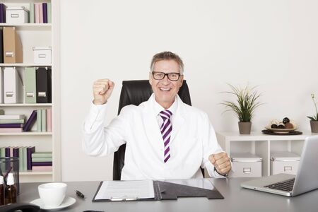 Jubilant middle-aged male doctor sitting cheering in his office punching the air with his fist as he celebrates a successful treatment, or promotionの写真素材
