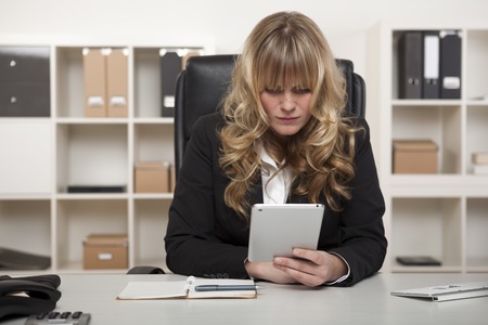Young businesswoman sitting at her desk in the office reading a message on her tablet at work, with a background of files on shelves.の写真素材