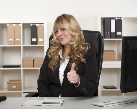 Attractive businesswoman with long curly blond hair sitting at her desk in the office giving a thumbs up gesture of approval and successの写真素材