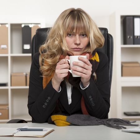 Young girl at work sitting at her desk in the office with a warm winter scarf around her neck enjoying a mug of hot coffeeの写真素材