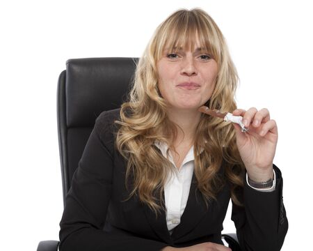 Beautiful young blond businesswoman eating a candy at her desk looking up at the camera with a mouthful and a guilty smileの写真素材