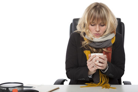 Cold businesswoman drinking hot coffee hunching over the hot liquid in a knitted winter scarf as she sits at her desk, on whiteの写真素材