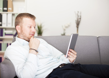 Thoughtful man reading his tablet computer as he relaxes on a couch with his hand to his chin and a pensive expressionの写真素材