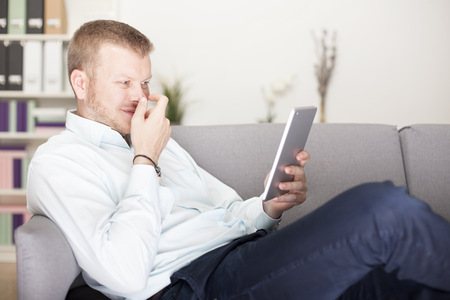 Man snickering to himself as he reads his tablet computer holding his hand in front of his face as he lies back comfortably on a sofa at homeの写真素材