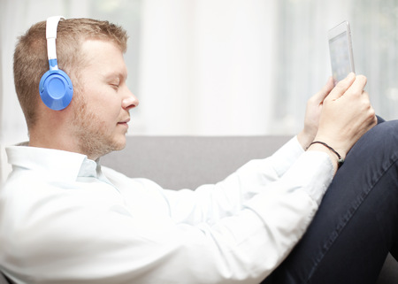 Man immersed in his music listening with closed eyes as he relaxes on a sofa with his feet up holding a tablet or MP3 storage device attached to his headphonesの写真素材