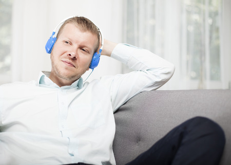 Young man smiling to himself as he listens to music on his headphones while relaxing on a sofa at home staring off into the air with a contented expressionの写真素材