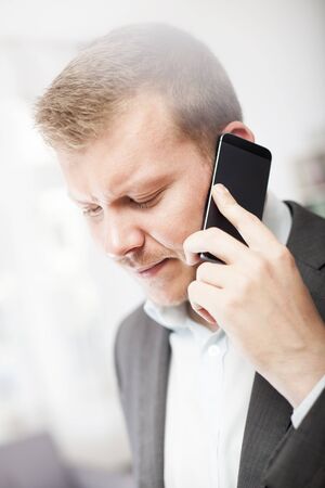 Businessman frowning at a call on his mobile as he stands listening to the conversation with downcast eyes, flare effect above his headの写真素材