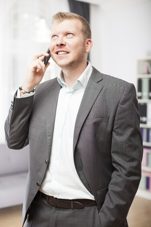 Stylish businessman in an open neck shirt and suit standing chatting on his mobile phone looking up into the air with a smileの写真素材
