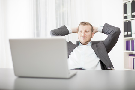 Successful confident young businessman sitting back in his chair at the office with his hands clasped behind his head and a satisfied expressionの写真素材