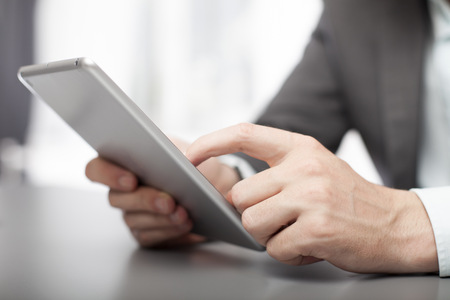 Businessman using his tablet in the office navigating the screen with his finger as he surfs the internet, close up view of his handの写真素材