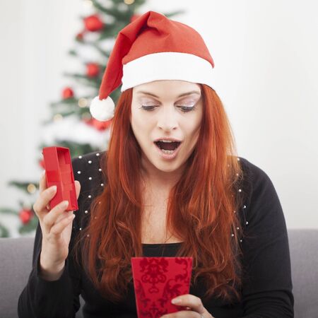 young red haired happy girl suprised open a christmas present, sitting in front of a firtree with lights and balls and wearing a santa hatの写真素材