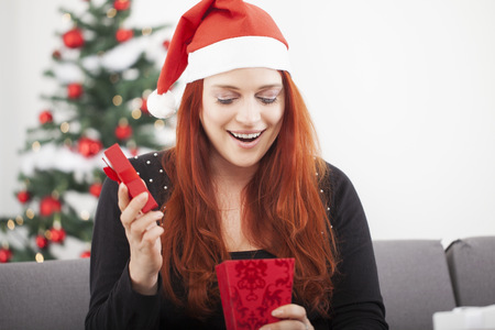 young red haired happy girl open a christmas present, sitting in front of a fir tree with lights and balls and wearing a santa hatの写真素材