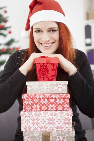 young red haired happy girl pose with christmas present, sitting in front of a fir tree with lights and balls and wearing a santa hatの写真素材