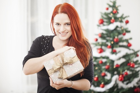 young red haired happy girl open a christmas present, sitting in front of a firtree with lights and balls and wearing a santa hatの写真素材