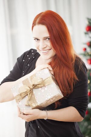 young red haired happy girl open a christmas present, sitting in front of a firtree with lights and balls and wearing a santa hatの写真素材